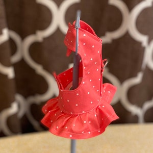 May include: A coral pink dog harness with white polka dots and a ruffled skirt. The harness is on a display stand.