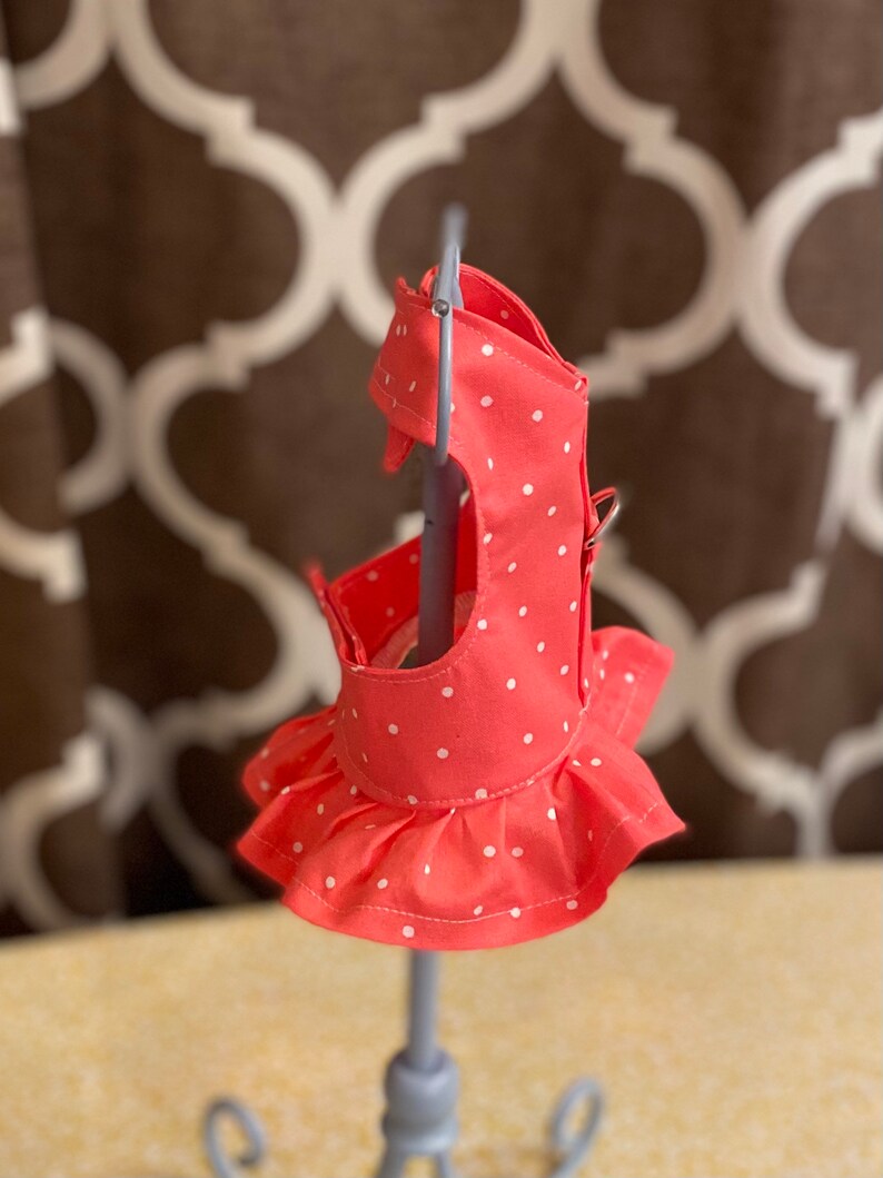 May include: A coral pink dog harness with white polka dots and a ruffled skirt. The harness is on a display stand.