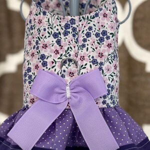 May include: A floral dog harness with a purple bow. The harness features a white background with pink and blue flowers. The skirt is purple with white polka dots and a dark purple trim. A metal ring is attached for leash attachment.