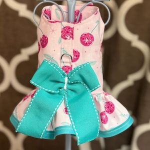 May include: A pink dog dress with a cherry print and a turquoise bow. The dress has a ruffled hem and a white trim.