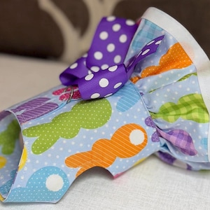 May include: A blue dog harness with a colorful Easter egg pattern and a purple polka dot bow. The harness has a white trim around the edges.