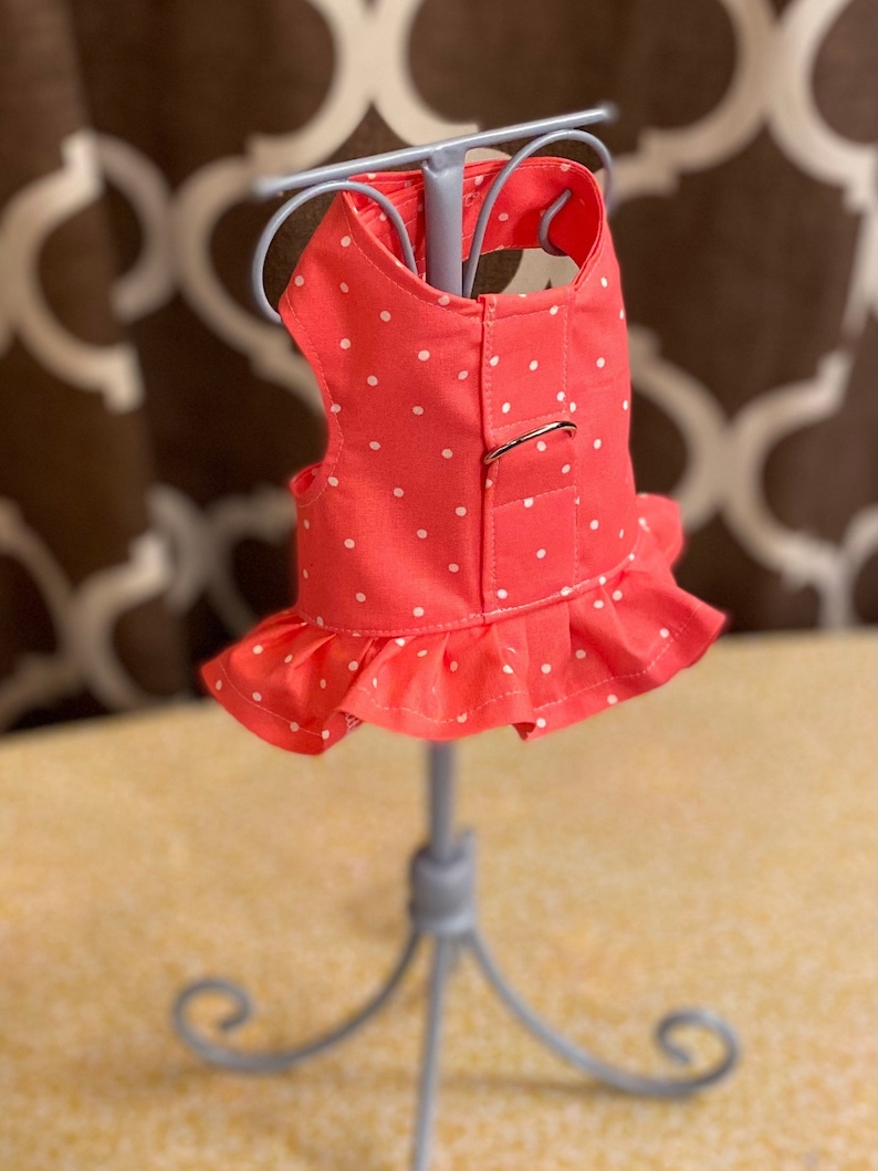 May include: A coral pink dog dress with white polka dots and a ruffle hem. The dress has a buckle closure on the back.