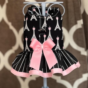 May include: Black dog dress with a pink bow and Eiffel Tower print. The dress features a black and white striped skirt with a pink trim. The Eiffel Tower print is white with pink accents.