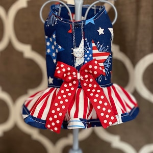 May include: A blue, red, and white dog dress with a red bow and white polka dots. The dress has a ruffled skirt and a festive Fourth of July theme.