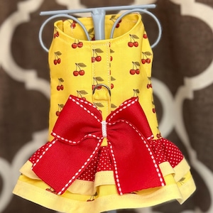 May include: A yellow dog harness with a cherry pattern, featuring a large red bow and a ruffled hem. The harness has a metal ring for leash attachment. The design includes a red and white polka dot pattern.