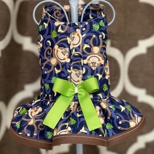 May include: A blue and brown dog dress with a monkey print and a green bow. The dress has a ruffled bottom and a brown trim.