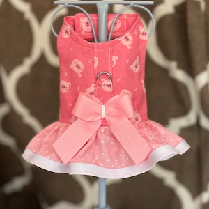 May include: A pink dog dress with a white trim and a pink bow. The dress has a pattern of pink pigs and hearts.