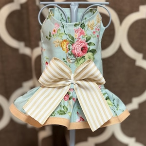 May include: A light blue dog dress with a floral pattern of pink and yellow roses. The dress features a large bow with tan and white stripes and a ruffled skirt with a tan trim. The dress is displayed on a light blue stand.