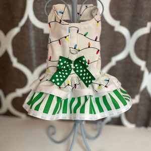 May include: A white dog dress with a green bow and a Christmas lights pattern. The dress has a green and white striped ruffle at the bottom.