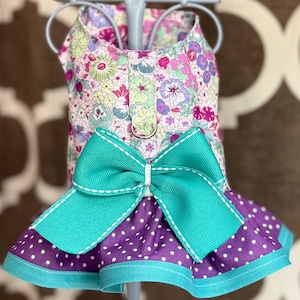 May include: A pet harness dress featuring a floral print bodice in shades of pink, green, and purple. It includes a large teal bow with white stitching at the waist, a purple skirt with white polka dots, and a teal hem. A metal D-ring is attached.