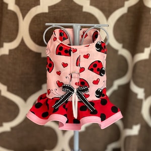 May include: Pink and red ladybug print dog dress with a black polka dot ruffle. The dress has a black ribbon bow at the back.