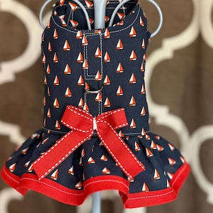 May include: Navy blue dog dress with a pattern of white and orange sailboats. The dress features a red bow and red trim along the hem. The dress is displayed on a light blue hanger.