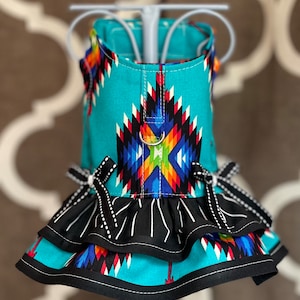 May include: A turquoise dog dress with a colorful geometric pattern and black ruffle trim. The dress has a black bow on each side and a silver ring on the back.