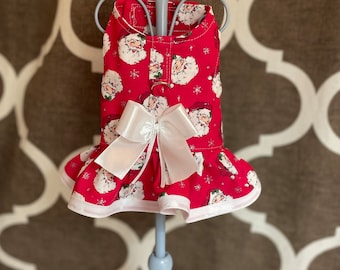 May include: Red and white dog dress with a Santa Claus print and a white bow. The dress has a ruffled hem and a white trim.