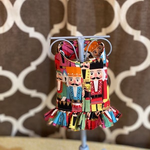 May include: A colorful dog dress with a Nutcracker print. The dress has a ruffled hem and is displayed on a white metal stand.