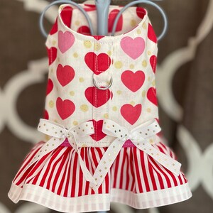 May include: A white dog harness with a red and pink heart pattern and gold glitter. The harness has a white ruffle with red stripes and gold glitter bows.
