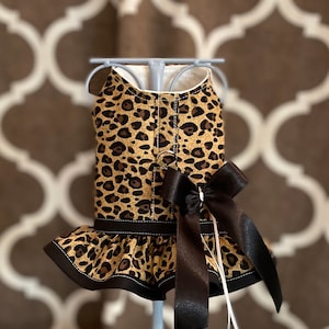 May include: A brown and black leopard print dog dress with a black satin bow and a white ruffle trim.