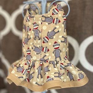 May include: A beige dog dress with a ruffle hem and a pattern of gray sock monkeys wearing red hats. The dress has a tan trim around the neckline and hem.