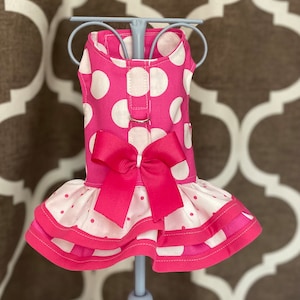May include: A pink and white polka dot dog dress with a pink bow and a ruffled skirt.