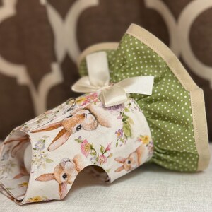 May include: An Easter-themed egg cozy featuring a bunny and floral print. The cozy is white with a bunny and floral pattern, and a green polka-dot ruffle with a cream-colored trim. A cream-colored bow adds a decorative touch.