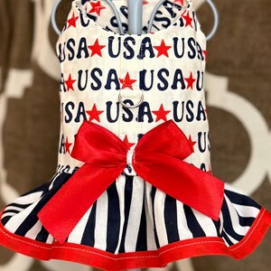 May include: A patriotic dog dress featuring a white background with the word "USA" and red stars. The dress has a red bow and a striped skirt with red trim. A metal ring is attached for a leash.