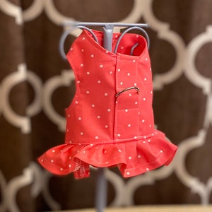 May include: A pink and white polka dot dog harness with a ruffle trim. The harness has a metal buckle and is designed for small dogs.