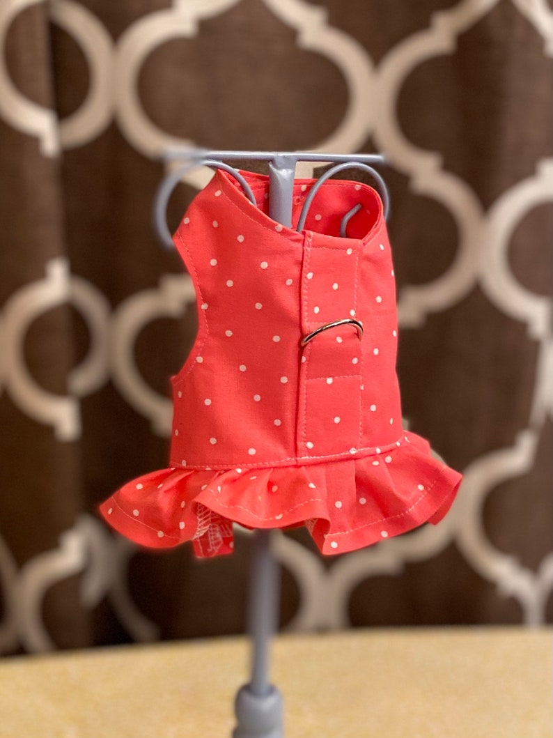 May include: A pink and white polka dot dog harness with a ruffle trim. The harness has a metal buckle and is designed for small dogs.