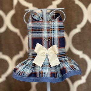 May include: A blue, orange, and navy plaid dog dress with a cream-colored bow. The dress has a ruffled skirt and a blue trim. The dress is displayed on a light blue mannequin.