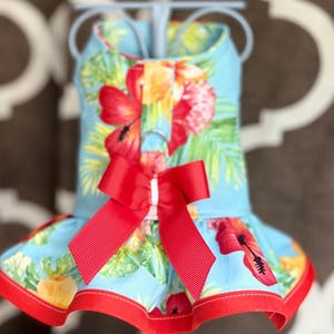 May include: A light blue pet harness dress featuring a vibrant tropical floral print with red hibiscus flowers and green palm leaves. The garment includes a solid red grosgrain ribbon bow at the center chest and a red fabric trim along the hemline.