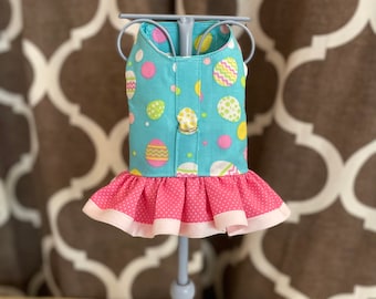 May include: A blue and pink dog dress with an Easter egg pattern. The dress has a ruffled skirt and a white trim.