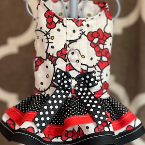 May include: A white dog harness with a Hello Kitty print and a black and white polka dot ruffle skirt. The harness has a red bow and a black buckle.
