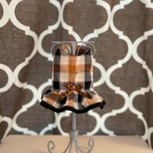 May include: A brown, black, and white plaid dog harness with a brown bow and brown heart-shaped accents. The harness is on a gray stand.