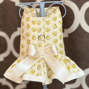 May include: A cream-colored dog dress featuring a pattern of yellow ducklings. The dress has a ruffled skirt, a satin bow, and a metal ring closure. The dress is designed for a small dog.