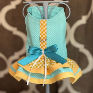 May include: A light blue dog dress with a yellow and white checkered pattern, a teal bow, and a yellow trim. The dress has a ruffle at the bottom.