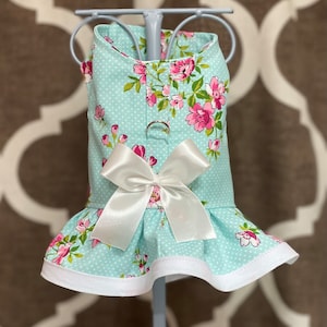 May include: A light blue floral dog dress with a white bow and white trim. The dress has a polka dot pattern and pink flowers.