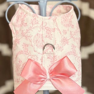May include: A pink and white floral patterned dog harness with a large pink bow. The harness has a metal ring for leash attachment and is displayed on a light blue stand. The background is a blurred pattern of brown and white.