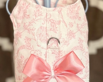 May include: A pink and white floral patterned dog harness with a large pink bow. The harness has a metal ring for leash attachment and is displayed on a light blue stand. The background is a blurred pattern of brown and white.