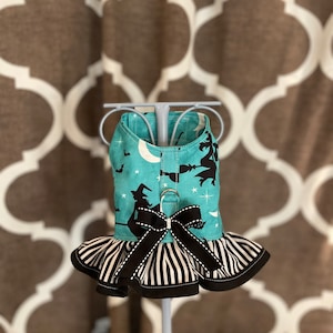 May include: A turquoise and black dog harness with a witch and moon print. The harness has a black and white striped ruffle and a black bow.
