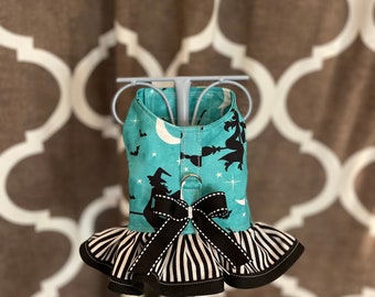 Puede incluir: Un arnés para perro turquesa y negro con estampado de bruja y luna. El arnés tiene un volante de rayas blancas y negras y un lazo negro.