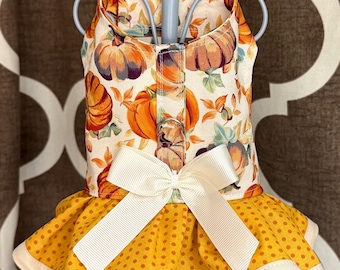 May include: A pet harness dress featuring a cream-colored bodice with an orange and brown pumpkin print. The skirt is mustard yellow with small brown polka dots and a cream-colored hem. A cream-colored ribbon bow is centered at the waist.