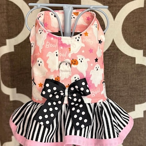 May include: A pet harness dress featuring a light pink fabric with a pattern of white ghosts, orange pumpkins, and stars. The bodice includes a black and white polka dot bow at the center, while the skirt has black and white vertical stripes.