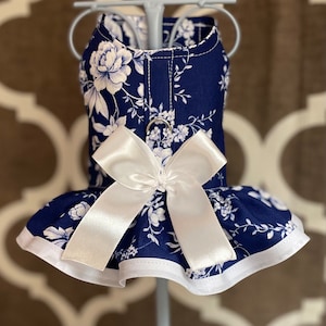 May include: A navy blue dog dress with a white floral print and a white satin bow. The dress has a ruffled hem and a white trim.