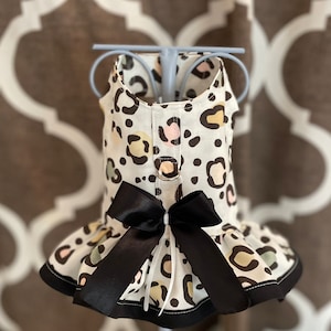 May include: A white dog dress with a black bow and a leopard print pattern. The dress has a ruffled skirt and a black trim.