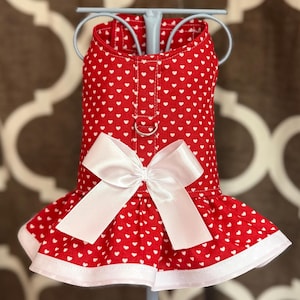 May include: Red dog dress with a white heart pattern and a white bow. The dress has a ruffled skirt with a white trim and a metal ring for leash attachment.