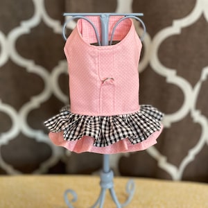 May include: Pink polka dot dog dress with a black and white gingham ruffle. The dress is displayed on a light blue mannequin. The background features a brown and white patterned design.