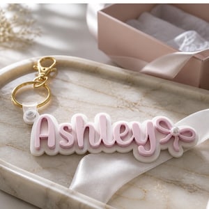 May include: A personalized keychain with the name "Ashley" in pink script lettering, outlined in white. The keychain features a gold-colored clasp and a decorative bow with a pearl accent. The design is set on a marble tray.