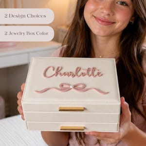 May include: A cream-colored jewelry box with two drawers and gold-colored handles. The box is personalized with the name "Charlotte" in pink script and a pink ribbon design. The image also shows text that reads "2 Design Choices" and "2 Jewelry Box Color."