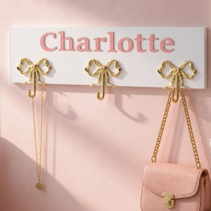 May include: A white rectangular wall-mounted hook rack featuring the name Charlotte in pink lettering. The rack includes three gold-coloured metal hooks shaped like bows. A gold chain necklace hangs from the left hook, and a pink purse hangs from the right.
