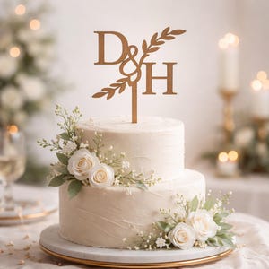 May include: A two-tiered white wedding cake decorated with white roses, greenery, and a gold cake topper featuring the letters "D & H". The cake sits on a gold-rimmed plate, with a blurred background of candles and flowers.