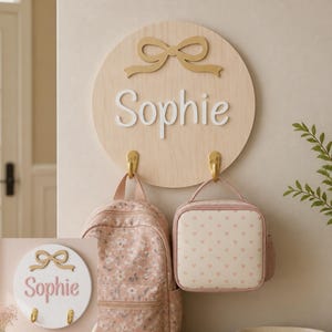 Peut inclure: Une plaque murale circulaire en bois clair avec un nœud doré en haut et le prénom Sophie en lettres 3D blanches. Deux crochets métalliques dorés sont fixés en bas, supportant un sac à dos à fleurs et un sac à déjeuner à motifs cœurs.